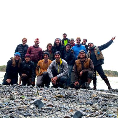 15 members of Islands Conservation Corps gather on a rocky beach with the ocean behind them. They are all smiling and wearing colorful outdoor clothing.