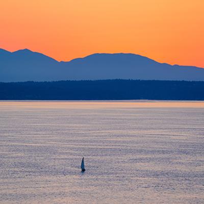 A lone sail boat on the waters of Puget Sound at dusk with an orange sky and silhouette of dark blue mountains in the background.