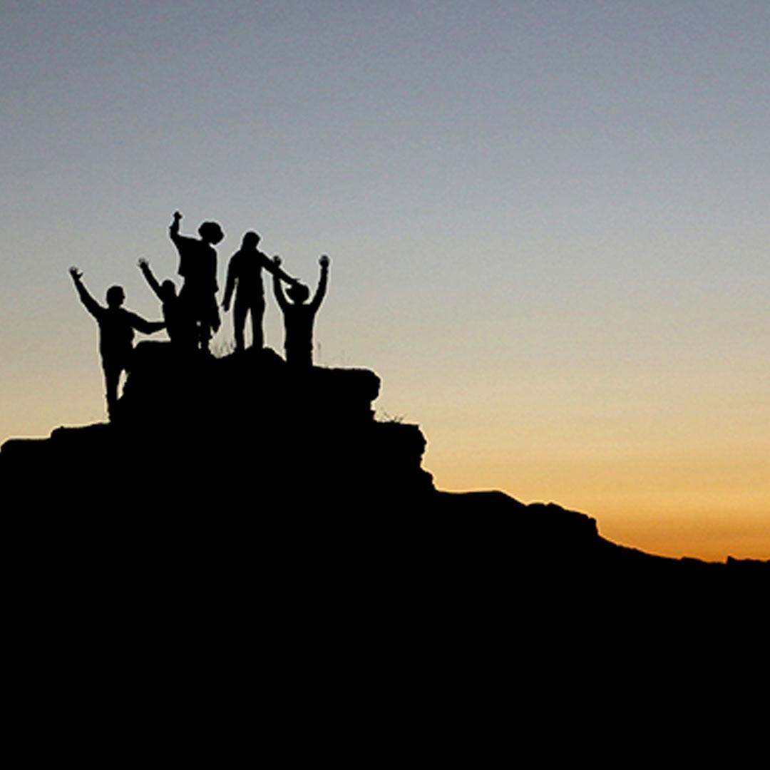 Silhouetted against a dusky sky, a small group of climbers stand with their arms outstretched on a rocky outcropping.